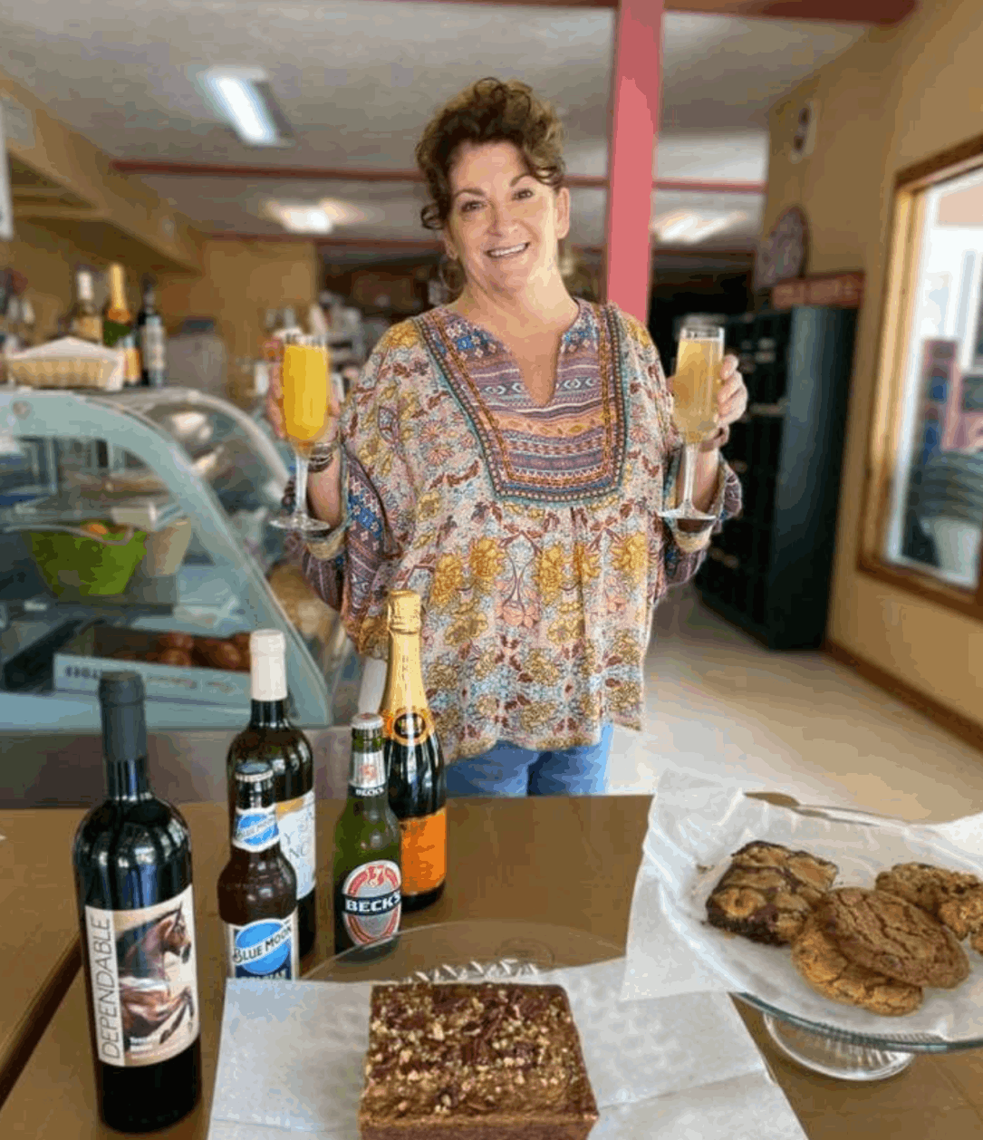 owner kelly harris hold wine galsses and a table with wine bottle and bake goods